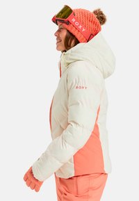 Aufgeblähte weiße und korallenfarbene Skijacke mit Kapuze, die einen Reißverschluss und Logodetails aufweist. Ergänzt durch eine passende Strickmütze und Skibrille.