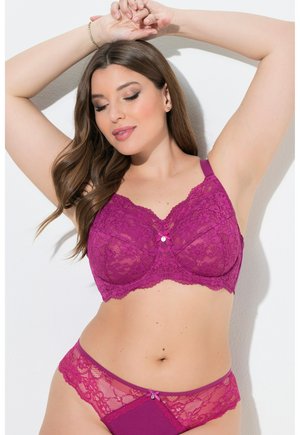 Frau mit langen braunen Haaren, die einen passenden lila Spitzen-BH und passende Unterwäsche trägt, posiert mit erhobenen Armen vor weißem Hintergrund.