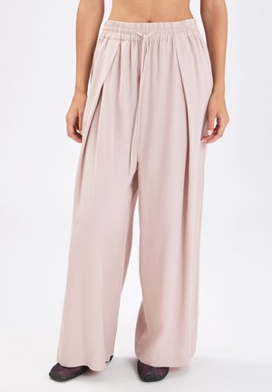 Donna che indossa pantaloni larghi a gamba larga di colore rosa pallido con cintura elastica e coulisse, abbinati a scarpe nere con punta, su sfondo bianco.