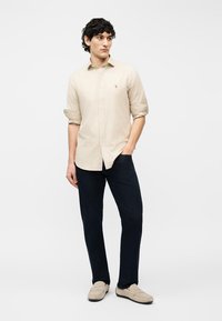 Νεαρός άνδρας με σγουρά μαύρα μαλλιά, ντυμένος με μπεζ ριγέ μακρυμάνικο πουκάμισο, σκούρο μπλε παντελόνι και μπεζ μοκασίνια, στέκεται με το ένα χέρι στην τσέπη.