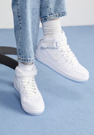Persoon die witte hoge sneakers met bandjes, witte geribbelde sokken en lichtblauwe jeans draagt op een lichtblauwe vloer.