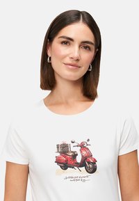 Frau mit braunen Haaren trägt ein weißes Shirt mit einem Grafik eines roten Rollers, der geschenkte Boxen im Leopardenmuster transportiert und darunter geschwungener Text.