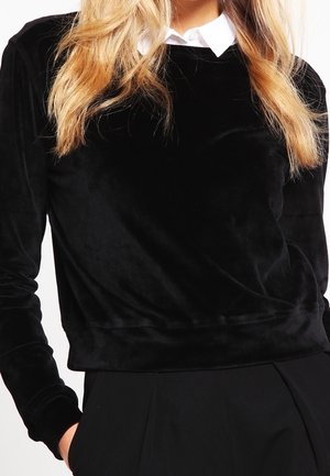 Femme portant un haut en velours noir à manches longues avec une chemise blanche colorée en dessous et un pantalon noir, les mains partiellement dans les poches.