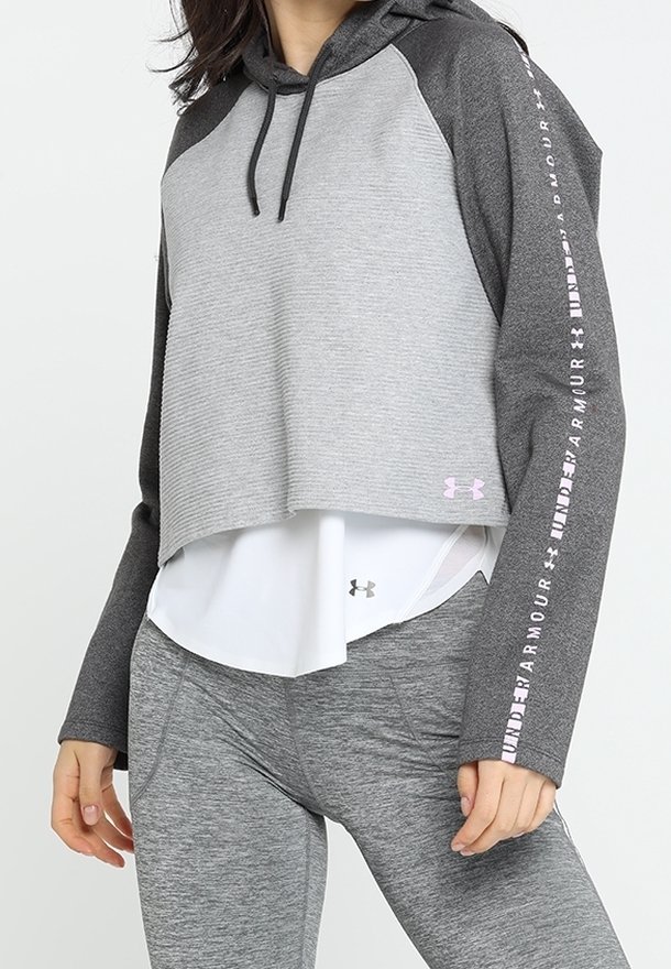 Femme portant un sweat à capuche court gris foncé avec le texte "Under Armour" sur les manches, chemise blanche superposée et leggings gris.