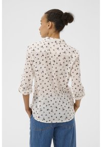 Chemise légère de couleur crème avec un motif floral. Comprend un col, des manches retroussables et un ourlet incurvé. Vue de dos visible.