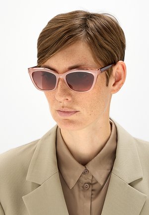 Persona con capelli castani corti che indossa grandi occhiali da sole rosa e una camicia beige con colletto sotto un blazer beige, guardando leggermente di lato.