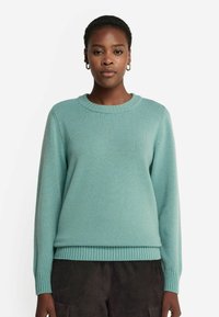 Jasnozielony sweter z okrągłym dekoltem, ściągaczami przy mankietach i na dole oraz miękką, fakturowaną powierzchnią. Noszony z ciemnymi spodniami.