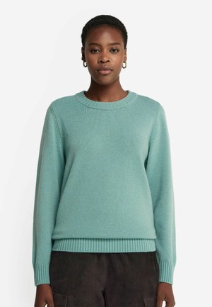 Hellblauer Pullover mit rundem Ausschnitt, gerippten Bündchen und Saum sowie einer weichen, strukturierten Oberfläche. Getragen mit dunklen Hosen.