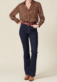Blus med leopardtryck och långa ärmar, i kombination med mörka jeans med utsvängda ben som har knappförslutning fram och ett brunt bälte med cirkulär spänne.