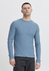 T-shirt à manches longues bleu en tissu texturé, doté d'un col rond et d'une coupe ajustée, associé à un pantalon sombre.