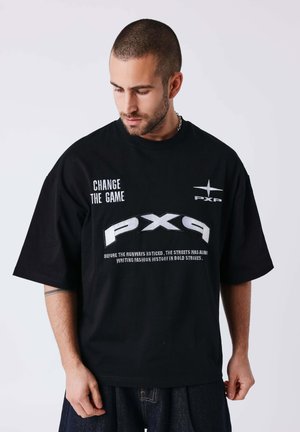Homme portant un T-shirt noir oversize avec un texte blanc "CHANGE THE GAME," grand "PX9," et un texte plus petit, regardant vers le bas sur sa gauche.
