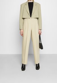 Blazer vert clair coupé sur un haut noir, associé à un pantalon beige taille haute. Le mannequin tient un petit sac à main noir et porte des bottines noires.