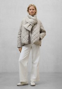 Gepatchte hellgraue Jacke mit Druckknöpfen, kombiniert mit einem passenden Schal, einem grauen Pullover und weiten weißen Hosen. Sneaker vervollständigen das Outfit.