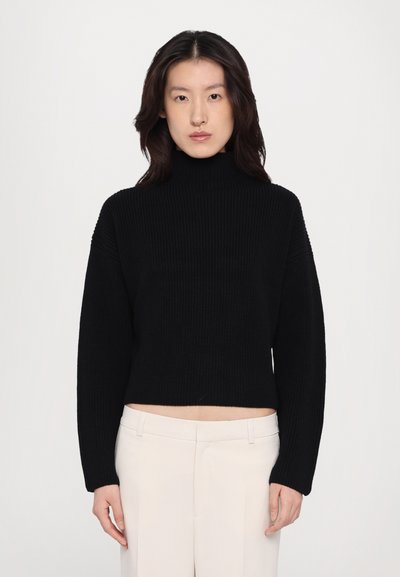 Giovane donna che indossa un maglione a collo alto a coste nero e pantaloni alla vita alta di colore bianco sporco, in piedi contro uno sfondo chiaro e semplice.