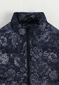 Veste matelassée florale avec col montant, présentant une base bleu foncé et un motif floral gris texturé, fermeture éclair à l'avant.