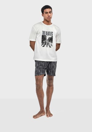 Jeune homme debout pieds nus portant un t-shirt blanc avec un motif graphique des Beatles et un short noir avec un motif répétitif du texte "The Beatles".