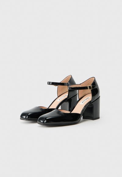 Par de sapatos Mary Jane de pele envernizada preta com saltos bloco, tiras no tornozelo e fechos de fivela dourada, sobre um fundo branco liso.