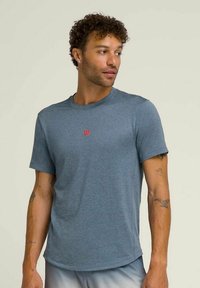 Camiseta deportiva gris de manga corta para hombre, confeccionada en una tela suave y elástica. Presenta un pequeño logo rojo en el pecho y un escote redondo.