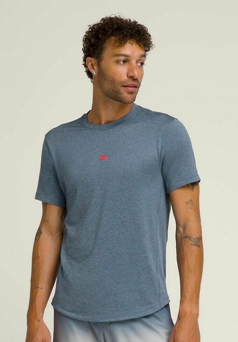 Camiseta deportiva gris de manga corta para hombre, confeccionada en una tela suave y elástica. Presenta un pequeño logo rojo en el pecho y un escote redondo.