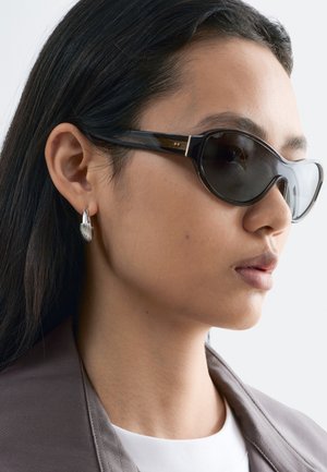 Mujer con cabello largo y oscuro, lleva gafas de sol tipo cat-eye de color oscuro, pendientes de aro plateados, una blusa blanca y una chaqueta gris, mirando hacia la derecha sobre un fondo blanco.