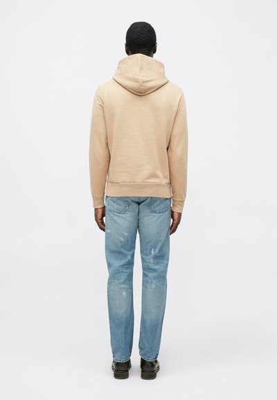 Μπεζ φούτερ με κουκούλα, με ριμπ μανσέτες και τελείωμα, συνδυασμένο με ανοιχτό μπλε τζιν με ταλαιπωρημένη όψη και μαύρα παπούτσια. Άσπρο φόντο.