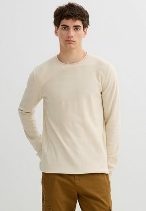 Joven con cabello rizado y oscuro, vistiendo un suéter crema de manga larga y pantalones marrones, sobre un fondo claro y liso.