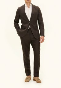 Completo in lino marrone con design aderente, giacca monopetto, revers a punta e due bottoni. Abbinato a una camicia chiara e mocassini beige.