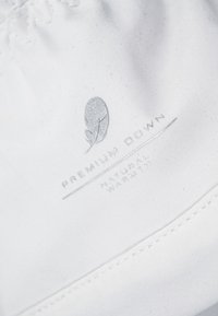 Étiquette en tissu blanc avec un texte argenté : "Plume Premium" et "Chaleur Naturelle", avec un logo en forme de feuille et un détail de bord cousu.