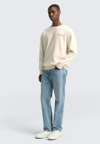Sweatshirt beige con scollo rotondo, maniche lunghe e un piccolo dettaglio testuale sul petto, abbinato a jeans dritti azzurri e sneakers bianche.
