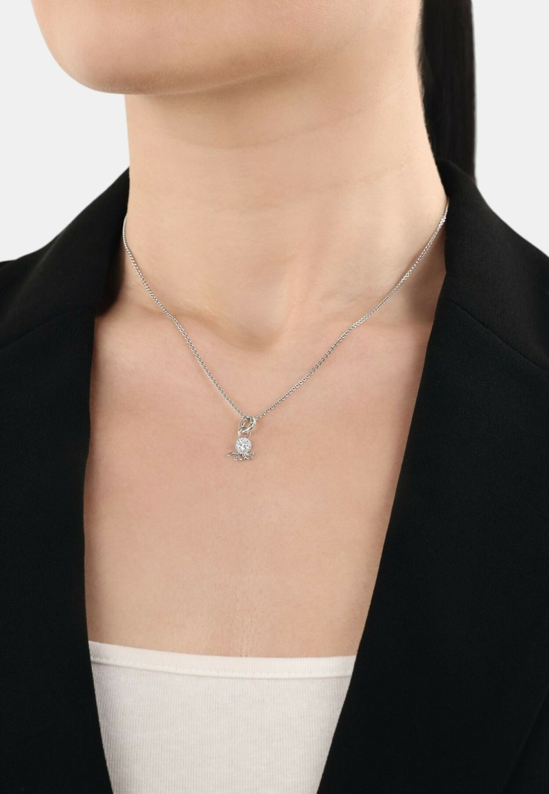 Collana d'argento con una delicata catena, caratterizzata da un pendente rotondo con un design floreale e piccoli dettagli, indossata sopra un blazer nero.