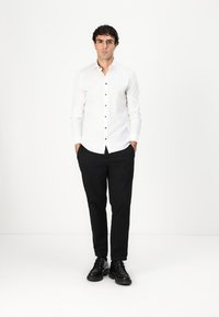 Hvit skjorte med mørke knapper, kombinert med svarte bukser og svarte sko. Antrekket har en tettsittende design og en uformell stil.