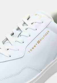 Nahaufnahme eines weißen Ledersneakers mit weißen Schnürsenkeln und dem goldenen Tommy Hilfiger-Logo auf der Seitenfläche.