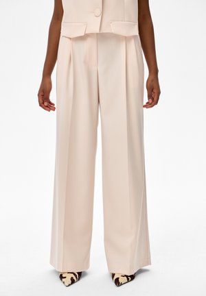 Kobieta ubrana w beżowe spodnie z wysokim stanem o szerokich nogawkach, w połączeniu z dopasowaną beżową kamizelką oraz szpiczastymi butami w dwukolorowy wzór zwierzęcy.