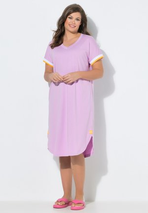 Kvinde i en knælang lavendelfarvet kjole med hvide og orange ærmekanter, der står barfodet i pink sandaler mod en hvid baggrund.