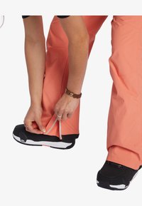 Waterdichte, fel koraalkleurige skibroek met een ritsmanchet, gecombineerd met zwarte snowboots. Focus op functionaliteit en ontwerp voor actieve kleding.