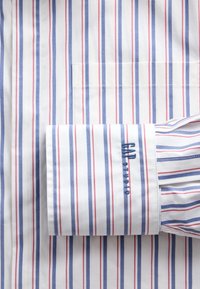 Chemise blanche à rayures verticales bleues et rouges, avec une poche et un poignet plié avec un texte brodé en bleu.