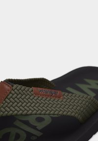 Πράσινο πλεκτό υφασμάτινο λουράκι με καφέ δερμάτινη ετικέτα που γράφει «Wrangler» σε μαύρο σανδάλι τύπου φλίπ-φλόπ με μερικώς ορατό λογότυπο.