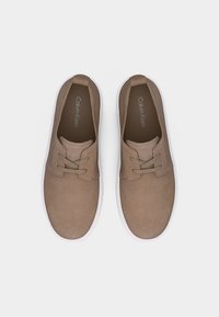 Ζευγάρι καφέ παπούτσια casual από καστόρι με κορδόνια και λευκές σόλες, απεικονιζόμενα από πάνω, με την εμπορική επωνυμία Calvin Klein στο εσωτερικό.