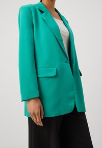 Blågrön kavaj med slag med utskärning, knappstängning med en knapp och framfickor, tillverkad av slät tyg, bärs över en vit topp och svarta byxor.