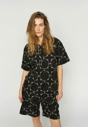 Naine, kellel on pikad lainelised juuksed, kannab musta lühikeste varrukatega särki ja sobivaid lühikesi pükse, millel on ümmargune viinapuu ja linnu muster.