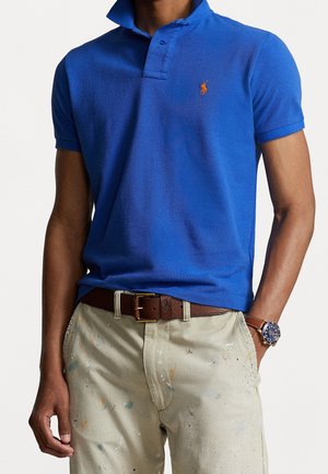 Mężczyzna w jasnoniebieskiej koszulce polo z krótkim rękawem i pomarańczowym logo, beżowych spodniach poplamionych farbą, brązowym skórzanym pasku oraz zegarku na rękę z brązowym paskiem.