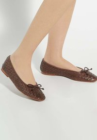 Brown woven ballet flats with a tied bow accent, flat sole, and pointy toe design, featuring a textured leather finish.
