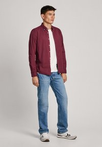Bordeaux langearmige shirt met knopen, gedragen over een wit t-shirt. Gecombineerd met lichtblauwe jeans en witte sneakers. Eenvoudig ontwerp, slim fit.