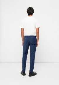 Mann mit dem Rücken zur Kamera, der ein weißes kurzärmliges Hemd, enganliegende blaue Hosen und schwarze Anzugschuhe vor einem schlichten weißen Hintergrund trägt.