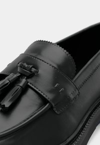 Black leather loafers with a glossy finish, textured band, and tassel accents. Features decorative stitching and a slightly rounded toe.