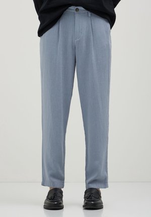 Hellblaue gerade geschnittene Hose, getragen mit schwarzen Quasten-Loafern bei einer Person, die vor einem einfarbigen Hintergrund steht.