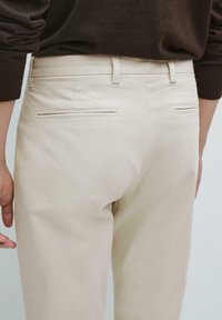 Beige bomuldsbukser med en tætsiddende pasform, der har to baglommer med stikninger og en slank talje. Glat tekstur.