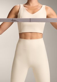 Svetlo bež rebrasti kropljeni top in legice z visokim pasom. Top ima V-izrez obliko. Spredaj je držana športna elastika z detajlom logotipa.