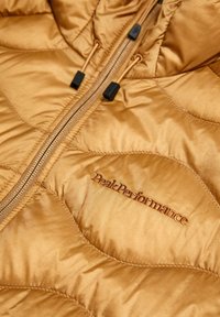 Gyldenbrun quiltet jakke med frontglidelås, snorstrikk og brodert "Peak Performance"-logo på brystet.