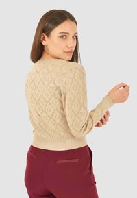 Donna con capelli castani lisci, che indossa un maglione lavorato a maglia beige e pantaloni bordeaux con vita alta, mentre guarda sopra la spalla.